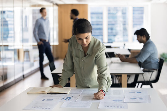 Focused Indian businesswoman analyst or strategist writing, making corrections, engaged in critical thinking, review financial report details, consider important information, do paperwork in office