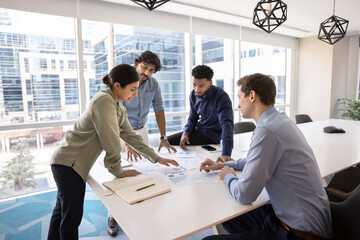 Indian professional woman expressing opinion, share solution, developing marketing strategy with diverse teammates gathered around conference table with heap of charts and graphs papers. Forecasting