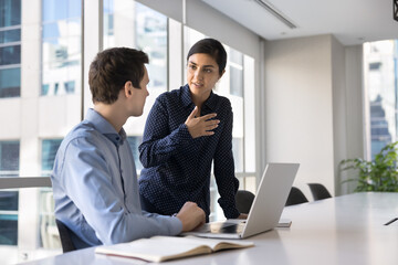 Young Indian woman and European man colleagues discussing joint task in office, met in modern conference room for opinion exchange, sharing solutions and point. Mentorship, apprenticeship, teamwork