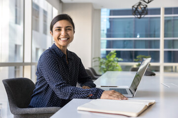Happy female corporate staff member posing for camera seated at table working on modern laptop in contemporary meeting room, exude confidence and competence. Professional Indian woman, office portrait