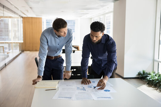 Two business professionals engaged in financial document analysis, reviewing reports, charts spread across table. African man points at specific data on sheet, explain insights or findings to teammate