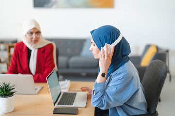 Two women in hijabs are working on laptops at a desk. One is listening to music with headphones. They are collaborating on a project in a bright, modern office.