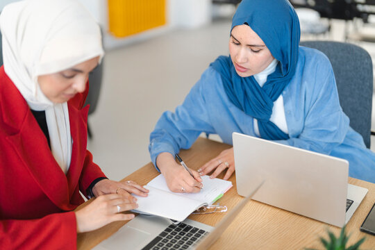 Two women in hijabs collaborate on a project. They're using laptops and notebooks to brainstorm ideas and take notes, aiming for success in their shared endeavor.