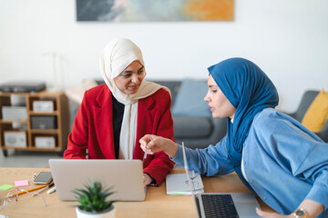 Two women in hijabs collaborate on a project. They're reviewing data on a laptop, brainstorming ideas, and working together to achieve a common goal.