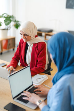 Two women in hijabs are collaborating on a project. They're using laptops and notebooks to brainstorm and work together, likely to achieve a shared goal in a professional setting.