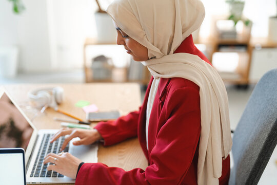 A woman wearing a hijab and red blazer is working on her laptop. She is focused on her work and typing efficiently.