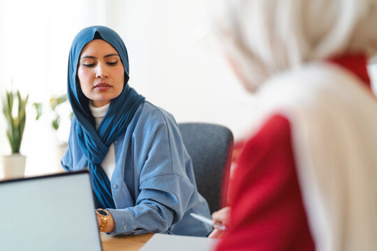 A woman in a blue hijab listens intently during a meeting. She's likely receiving feedback or discussing a project with a colleague, focusing on the details at hand.