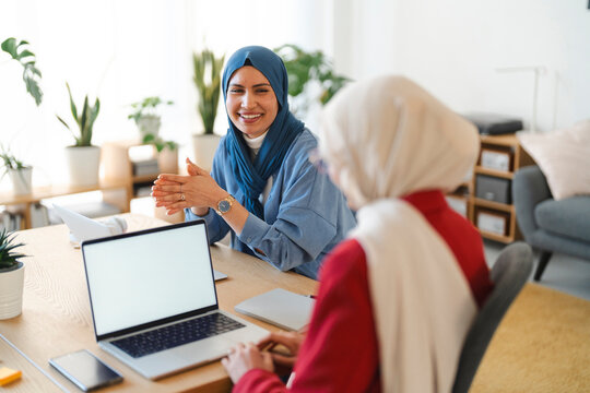 Two women in hijabs are collaborating on a project. They're working together at a desk with laptops, likely brainstorming or reviewing progress, fostering teamwork and innovation.