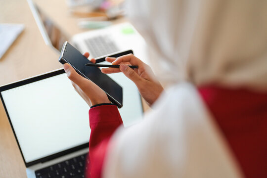 A woman in a hijab uses a stylus on her phone. She's likely working or studying, using technology to enhance her productivity.