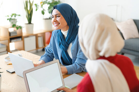 Two women are collaborating on a project. They're using laptops at a desk, likely brainstorming or working together on a presentation. The woman in blue seems happy with their progress.