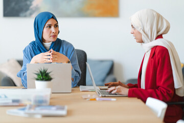 Two businesswomen in hijabs collaborate on a project. They're using laptops to brainstorm ideas and strategize for their company's growth.