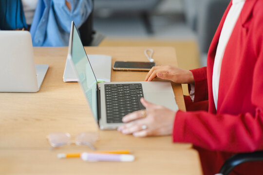 A woman in a red blazer works on her laptop at a wooden desk. She's likely preparing for a meeting or working on a project with colleagues nearby.