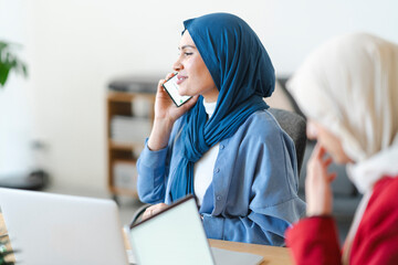 A woman in a blue hijab is on a call while working at a desk with a laptop. She's likely discussing work or personal matters, staying connected while managing her tasks.