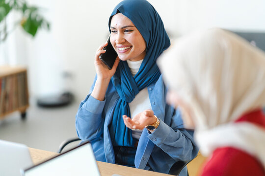 A woman in a blue hijab smiles while talking on her phone at work. She's likely discussing a project or client, as she gestures with her hand while sitting at her desk.