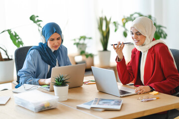 Two women in hijabs are working at a shared desk. One is on her phone, possibly dictating a message, while the other focuses on her laptop, maybe collaborating on a project.