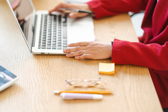 A person in a red blazer is working on a laptop. They are likely working on a project or task that requires the use of a computer. There are also glasses, pens, and a sticky note on the desk.