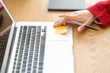 A person in a red blazer is using a pen to point at a sticky note on a laptop. The note reminds them of a meeting to stay organized and on schedule.