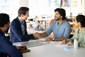 Two multiethnic business partners shaking hands sitting at desk with diverse companions, conclude...