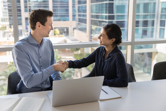 Happy colleagues shaking hands, finish formal meeting successfully, establish partnership, making commercial deal seated at table with laptop in contemporary corporate city office with skyscraper view