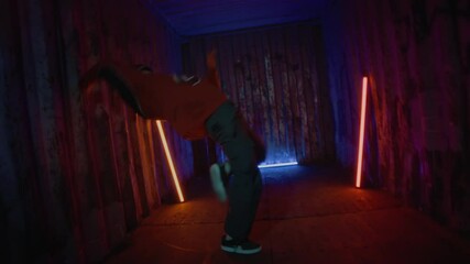 Full shot of young talented Caucasian b-boy performing breakdance indoors under blue and orange ambient light showing excellent footwork and creativity