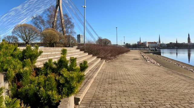 Scenic view of the Vansu Bridge from Kipsala Island in Riga, Latvia, with the cityscape in the background, featuring a paved promenade, lush greenery, and concrete steps, on a bright sunny day