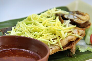 Fried star pomfret fish served with spicy mango sambal on a white plate. Traditional Indonesian seafood dish with a fresh, tangy, and savory flavor combination.