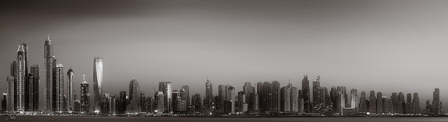 Fototapeta premium Monochrome city skyline with tall skyscrapers reflecting on calm water under a cloudy sky, Dubai Marina bay UAE