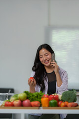 Young asian nutritionist talking on the phone and holding a tomato with various vegetables lying on...