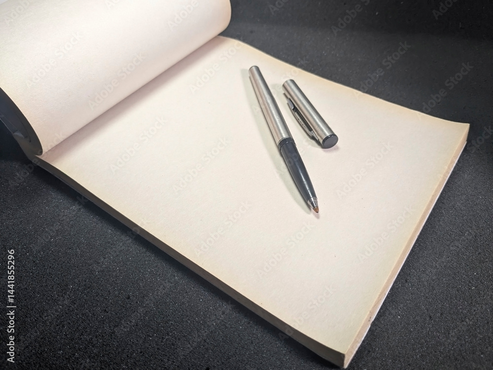 Poster close-up shot of a silver pen resting on an open notepad, capturing the concepts of writing, plannin
