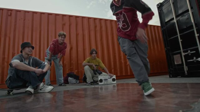 Handheld shot of confident Caucasian gen Z b-boy in urbanwear spinning his body while performing breathtaking breakdance moves, practicing outdoors among peers watching him