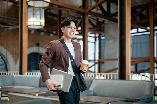 Brown suit man with glasses walking while holding a coffee cup with laptop and notebook in the cafe.