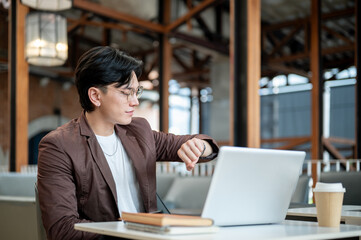 Fototapeta premium Brown suit asian man with glasses is looking at his wristwatch while sitting in cafe or coffee shop.