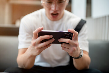 Asian man wearing glasses in white shirt is holding phone horizontally while sitting in the cafe.
