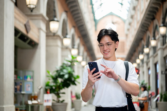 Asian man with glasses in white tee is smiling while looking at his phone in shopping mall or market