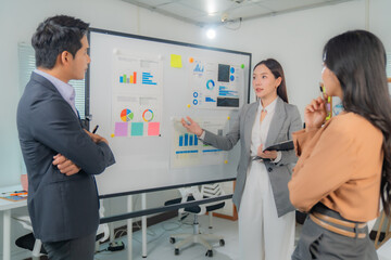 Fototapeta premium Three Asian business professionals engaging in a strategic discussion during a meeting in a modern office, utilizing a whiteboard filled with insightful charts and graphs