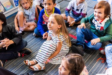 Children sitting on a carpet in a classroom, listening attentively. Diverse group of kids, including a blonde girl in a striped dress, engaged in learning. Young diverse children learning in classroom