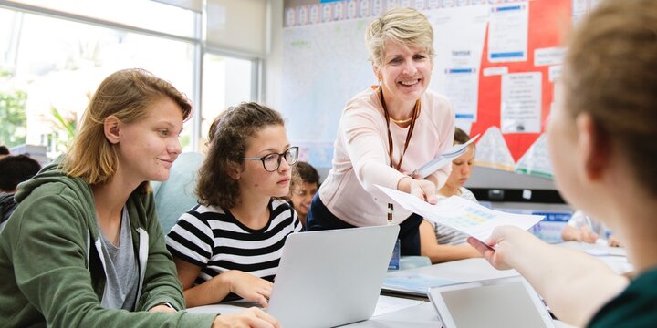 Classroom with diverse students and teacher, work on school group project. Diverse students work on laptop in classroom. Teacher engages with students, group project collaboration in school classroom