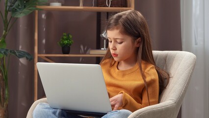 Young girl child spending evening time at home, girl sits in chair in the living room and working...