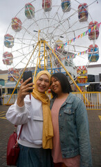Two Asian Woman Take Selfie At Amusement Park With Ferris Wheel On The Background