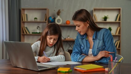 Obraz premium Young child girl studying at home, small girl sits at desk and attends school class online on laptop, doing homework, mother helping and controlling process.