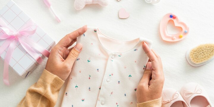 Hands holding a baby onesie with pastel patterns. Baby gift, baby clothes, and baby shower items like shoes and a brush. Soft, pastel, and baby-themed decor. Girl baby shower background.