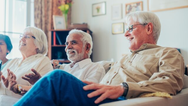 Group of diverse elderly friends sitting on a couch, laughing and enjoying each other's company in a cozy living room setting. Diverse old people talking and sitting on a sofa.