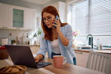 Mid adult woman talking on cell phone while working on laptop at home.