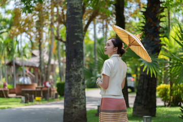 Fototapeta premium Young woman wearing traditional thai dress walking in tropical garden holding parasol looking away from camera, enjoying sunny weather in exotic location