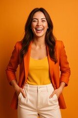 A joyful woman with long brown hair, standing in front of a vibrant, solid orange background, wearing a stylish orange blazer over a yellow top, paired with white high-waisted trousers.