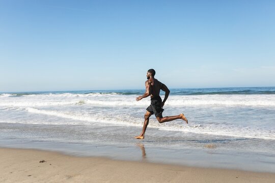 Man running on beach, ocean waves in background. Athletic man, beach running, enjoying the ocean. Fitness on the beach, man running by the ocean. African American man doing exercise workout.