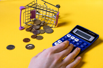 Hand managing expenses on blue calculator near scattered coins and fallen shopping cart on yellow