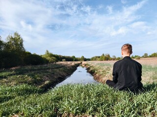 young guy is sitting alone in a meadow in early spring, against the background of a calm narrow...