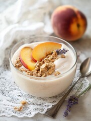 Bowl of yogurt with granola and sliced peaches on top. the bowl is placed on a white lace tablecloth with a spoon resting on the side.