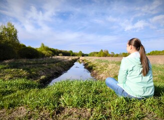 young woman is sitting alone in a meadow in early spring, against the background of a calm narrow...